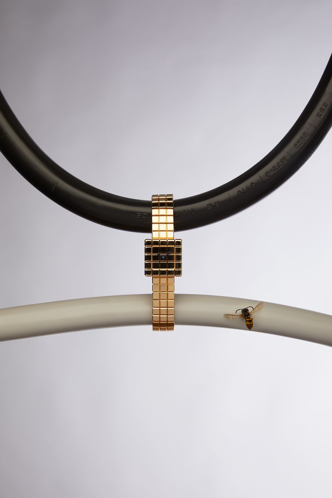Gold wristwatch suspended between curved black and white pipes in a studio shot with a bee alighting on the white pipe.