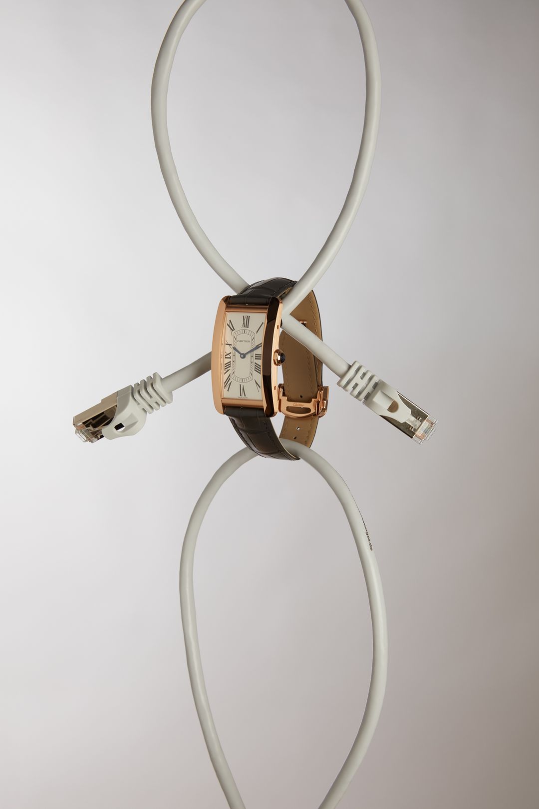 Rose gold wristwatch with a brown leather strap suspended by two white network cables on a light gray studio background.