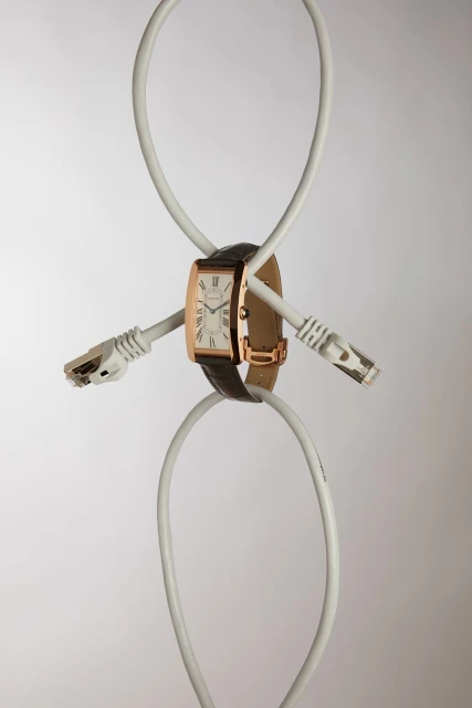 Rose gold wristwatch with a brown leather strap suspended by two white network cables on a light gray studio background.