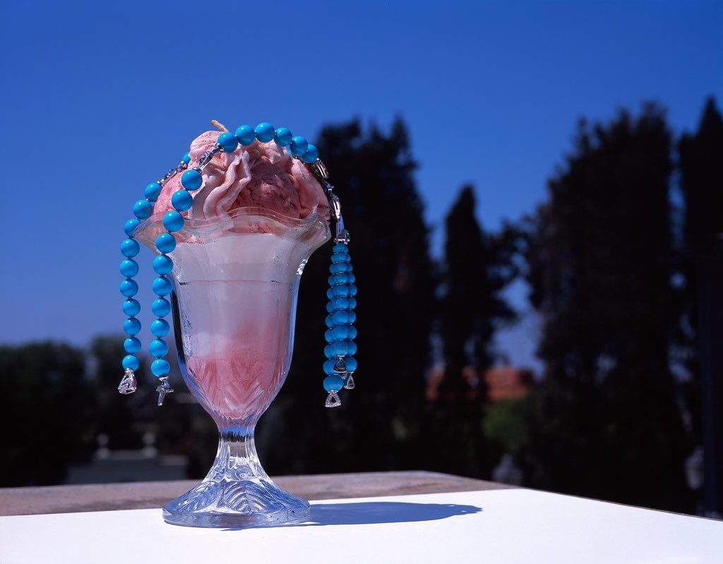 Crystal dessert glass filled with a light pink dessert and a blue beaded necklace in front of trees against a bright blue sky.