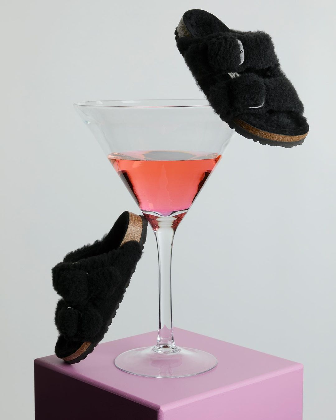 Fuzzy black slide sandals, cocktail glass with pink liquid, and pink cube create a surreal still life product shot on white.