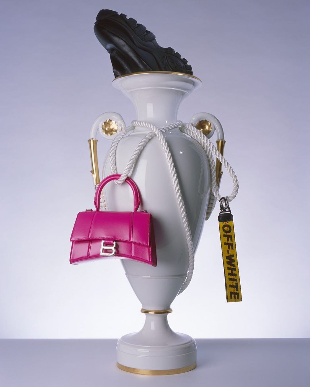 Still life with a black shoe on a white urn, a pink purse, and a yellow Off-White tag against a gradient blue background.