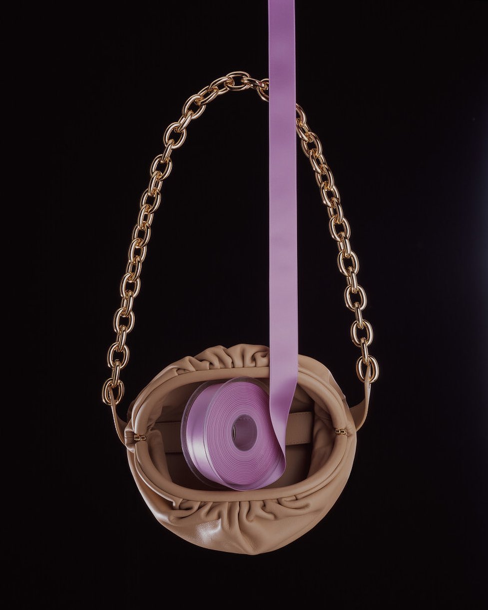 Elegant beige purse with gold chain handle and a roll of lilac ribbon cascading down against a stark black backdrop.