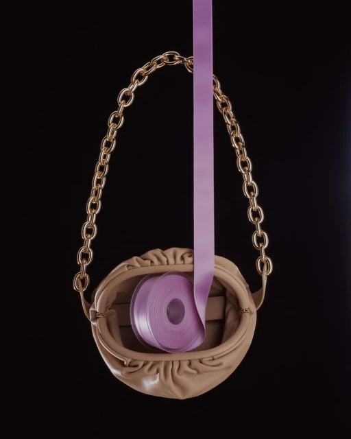 Elegant beige purse with gold chain handle and a roll of lilac ribbon cascading down against a stark black backdrop.