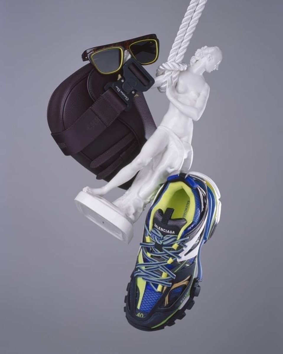 Fashion still life featuring sunglasses, brown purse, a white sculpture, a Balenciaga sneaker, and a white rope on a grey backdrop.