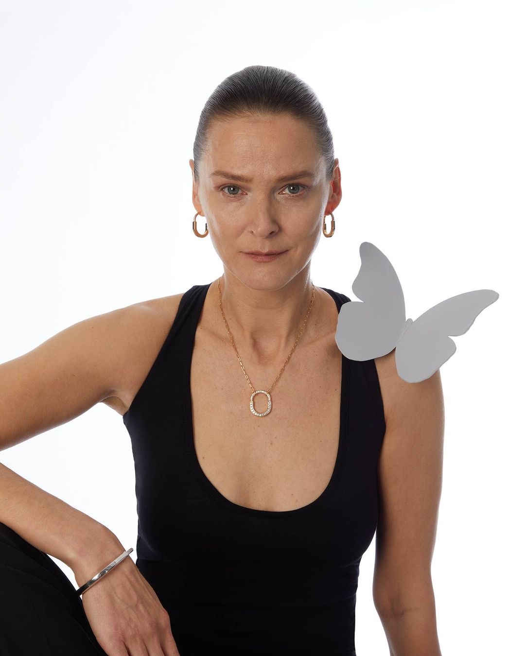 Portrait of a woman with hoop earrings and a gold necklace, black tank top, and a butterfly prop against a white backdrop.