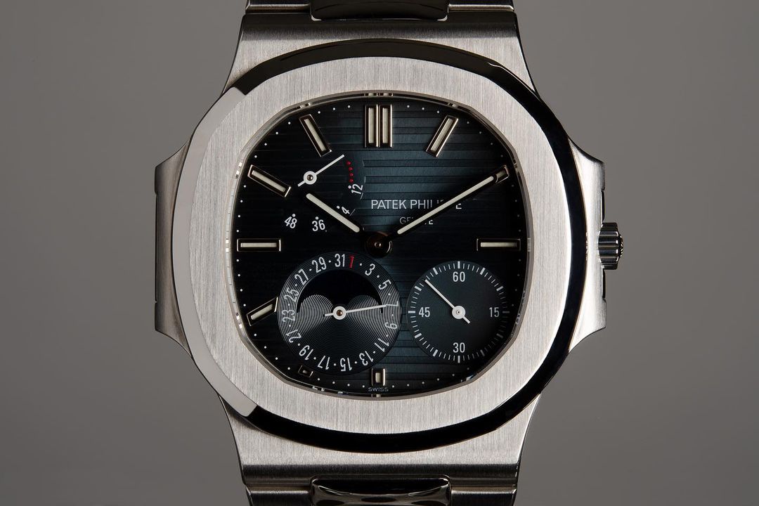Close-up studio shot of a Patek Philippe wristwatch with a silver band and a dark dial displaying time and moon phases.