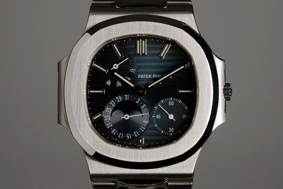 Close-up studio shot of a Patek Philippe wristwatch with a silver band and a dark dial displaying time and moon phases.