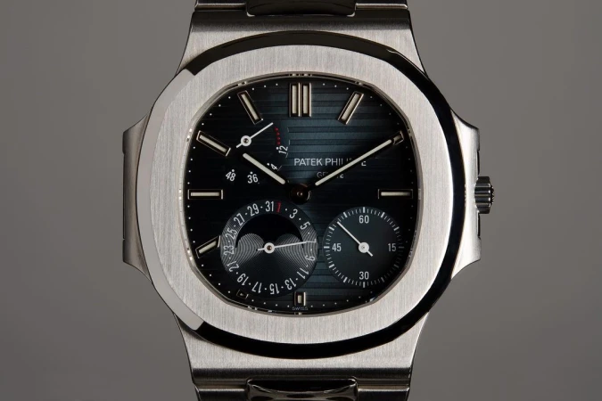 Close-up studio shot of a Patek Philippe wristwatch with a silver band and a dark dial displaying time and moon phases.
