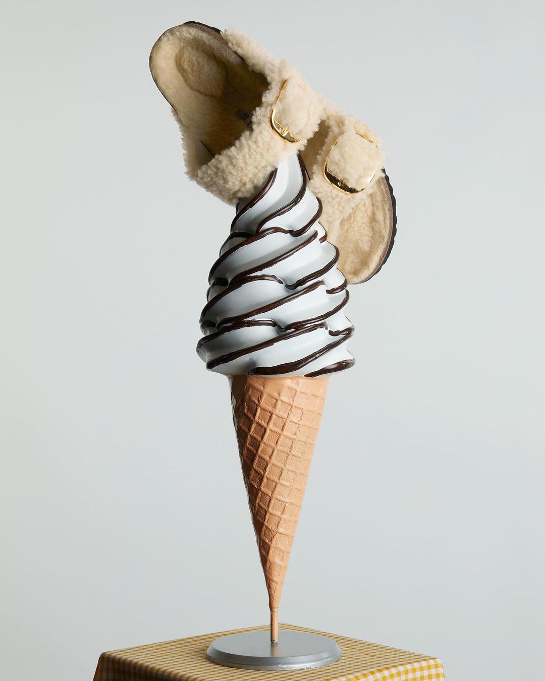 Conceptual fashion still life: a slip-on shoe perched atop a cone of white ice cream with chocolate drizzle on a checkered surface.