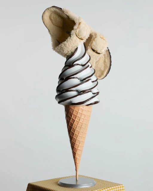 Conceptual fashion still life: a slip-on shoe perched atop a cone of white ice cream with chocolate drizzle on a checkered surface.