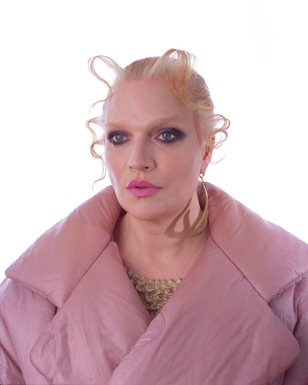 Portrait of a blonde woman with pink lipstick wearing a pink puffer coat, set against a bright white background in a studio photograph.