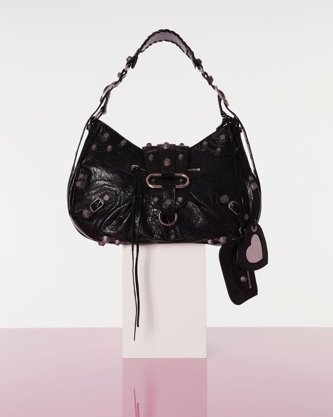 Black Balenciaga handbag with silver hardware, including studs and buckles, resting on a white block against a white to pink gradient backdrop.