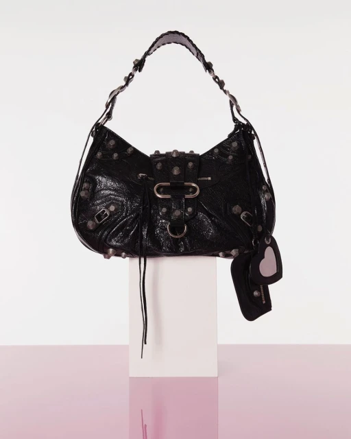Black Balenciaga handbag with silver hardware, including studs and buckles, resting on a white block against a white to pink gradient backdrop.