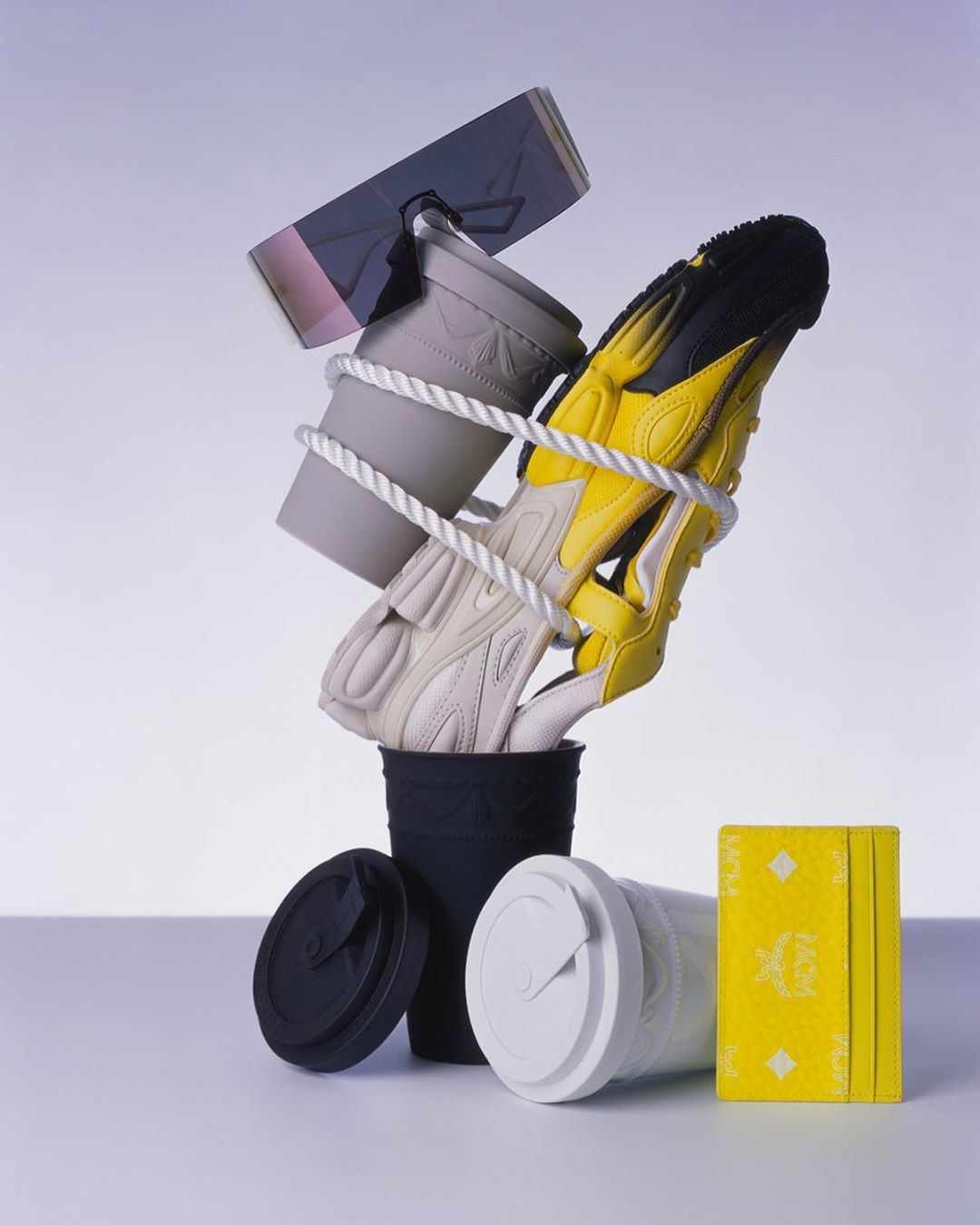 Still life featuring sneakers and yellow sandals, tied with white rope, resting in black and white cups with lids and a yellow book.