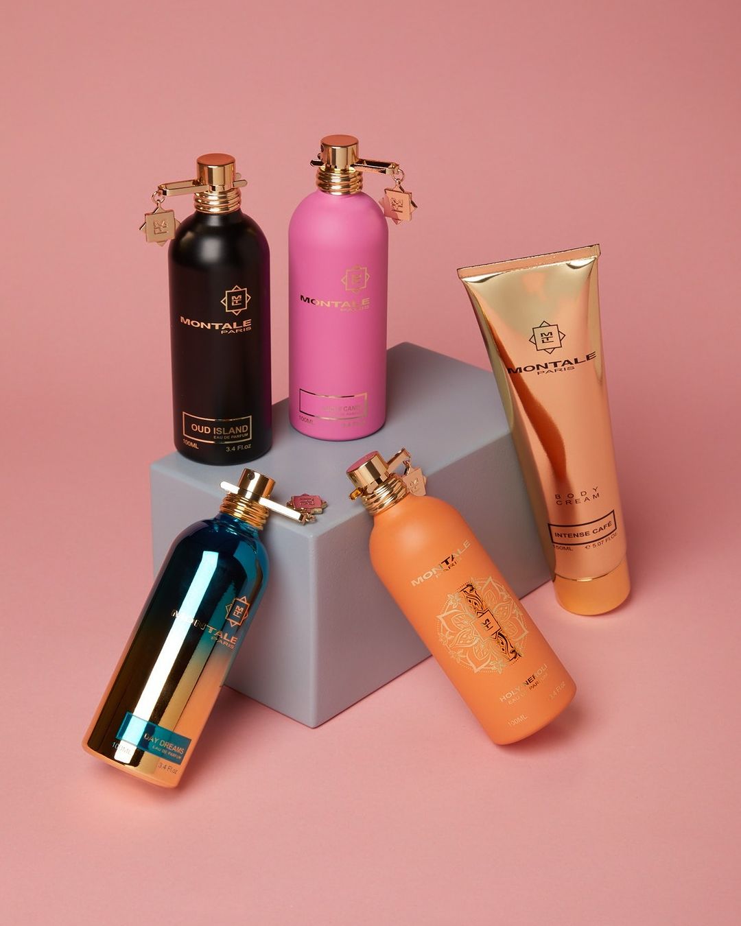 Still life product shot featuring five Montale perfume and body care bottles in black, pink, blue, and orange against a pink background.