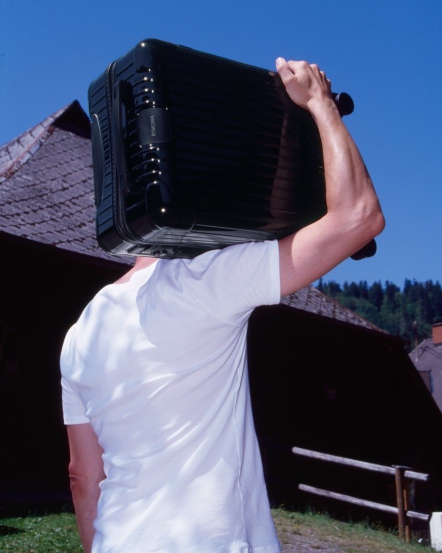 Adult carries a black hard shell suitcase on their head outdoors, wearing a white t-shirt, with a building and trees in the background.