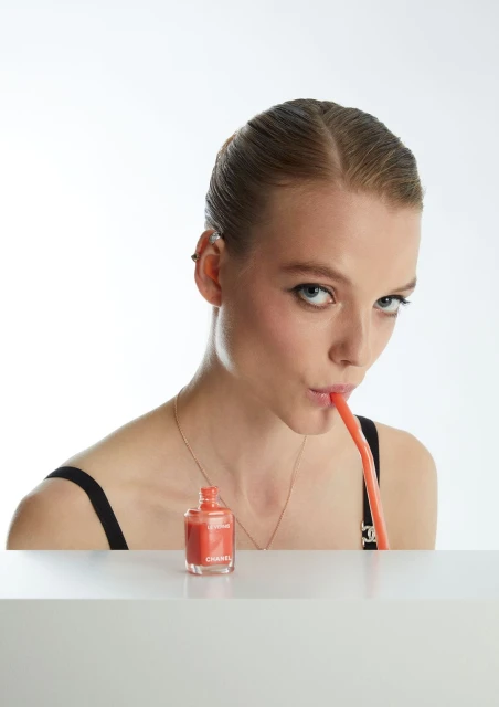 Woman poses with coral nail polish bottle and straw. Beauty product photography with a light backdrop. Studio shot for cosmetics.