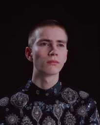 Close-up portrait of a young man with a shaved head wearing a dark patterned top against a black background.