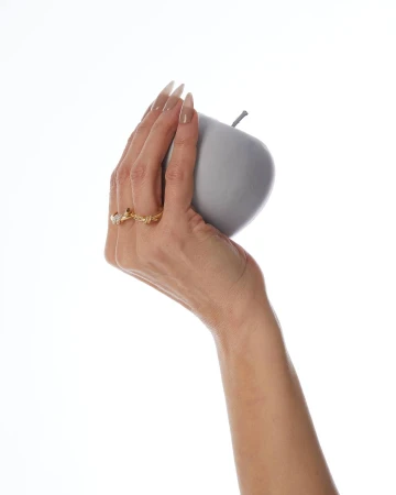 Close-up of a hand wearing gold rings holding a smooth grey artificial apple against a white background