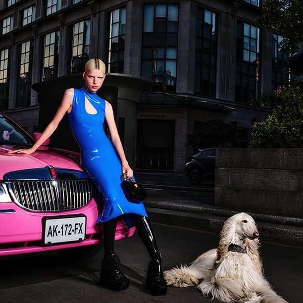 A woman poses next to a pink car with a dog in an urban setting. The woman is wearing a blue dress and black boots.