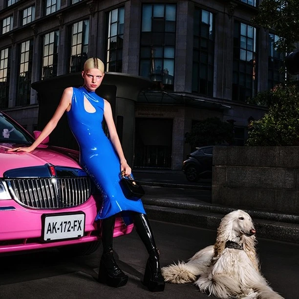 A woman poses next to a pink car with a dog in an urban setting. The woman is wearing a blue dress and black boots.