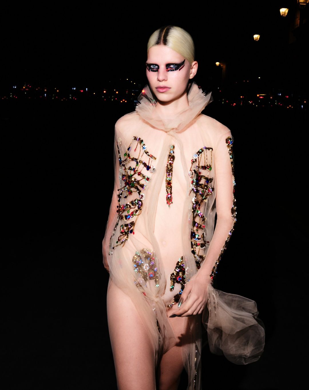 A person stands outdoors at night wearing a sheer beige dress with colorful embellishments and heavy eye makeup under streetlights.