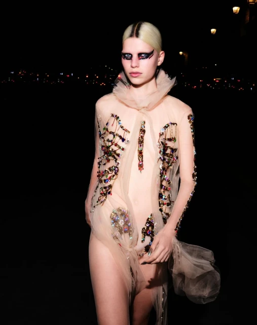 A person stands outdoors at night wearing a sheer beige dress with colorful embellishments and heavy eye makeup under streetlights.