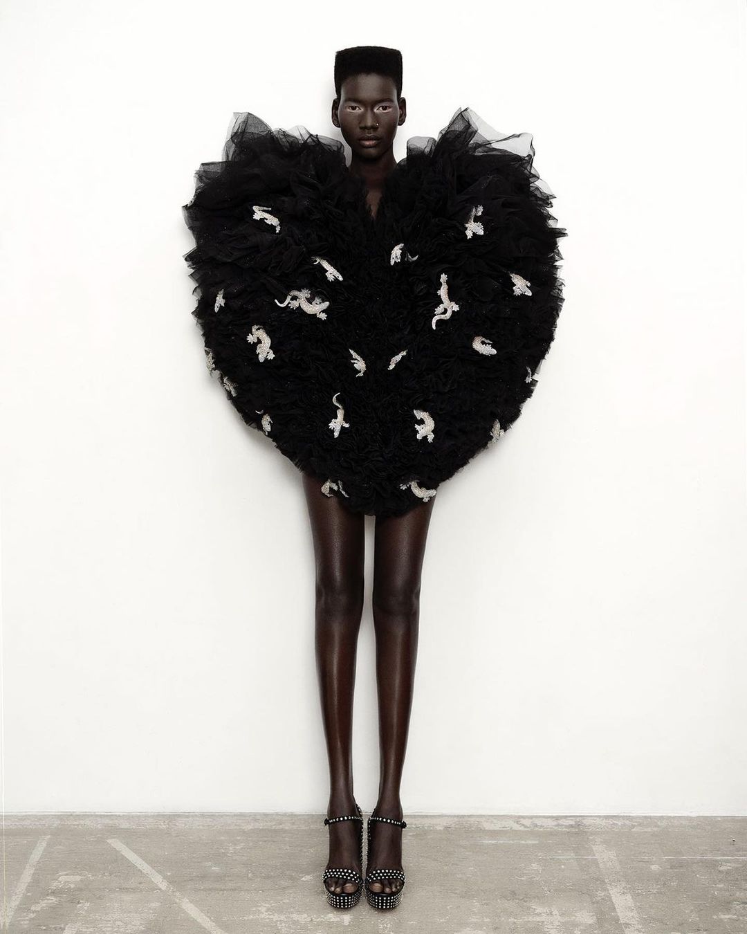 Fashion editorial shot of a Black woman in a large, ornate black dress with white designs, standing against a white wall in platform heels.