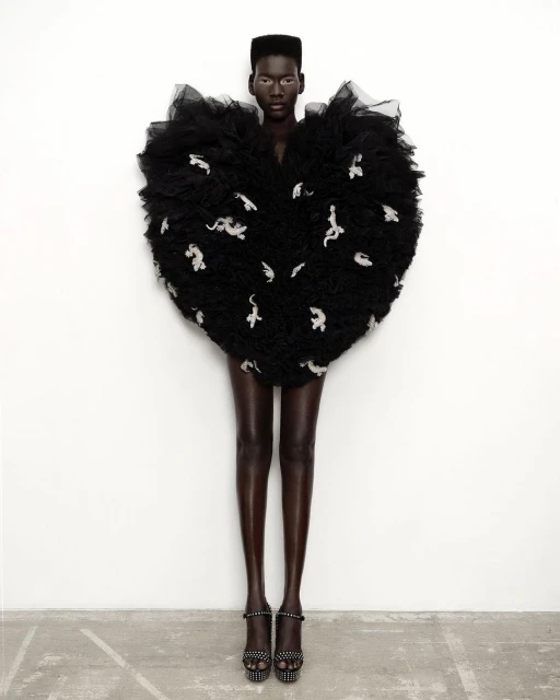 Fashion editorial shot of a Black woman in a large, ornate black dress with white designs, standing against a white wall in platform heels.