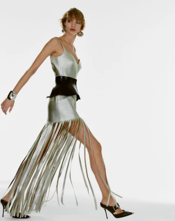 Model in silver fringe dress with black belt walking against a plain white background