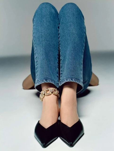 Close-up studio shot of a person sitting with their legs in the air, wearing jeans, black heels and a gold ankle bracelet.