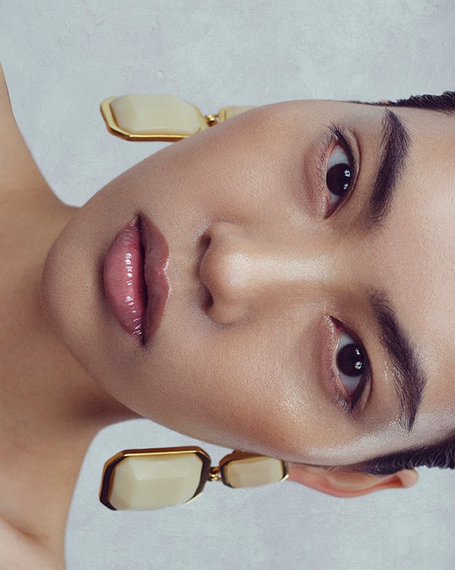 Close-up beauty shot of a woman with glossy lips and modern ivory and gold earrings against a soft, light grey background.