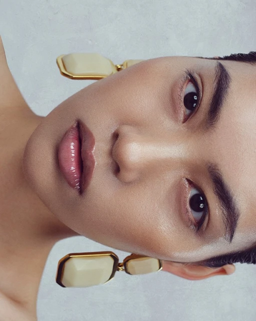 Close-up beauty shot of a woman with glossy lips and modern ivory and gold earrings against a soft, light grey background.