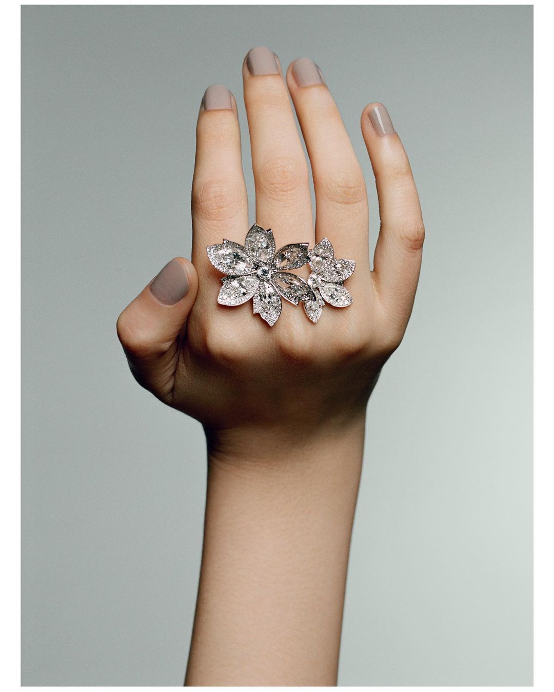 Close-up of hand wearing a large floral-shaped diamond ring. The ring features multiple petal-shaped diamonds on a light grey background.