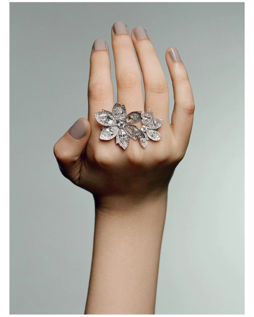 Close-up of hand wearing a large floral-shaped diamond ring. The ring features multiple petal-shaped diamonds on a light grey background.