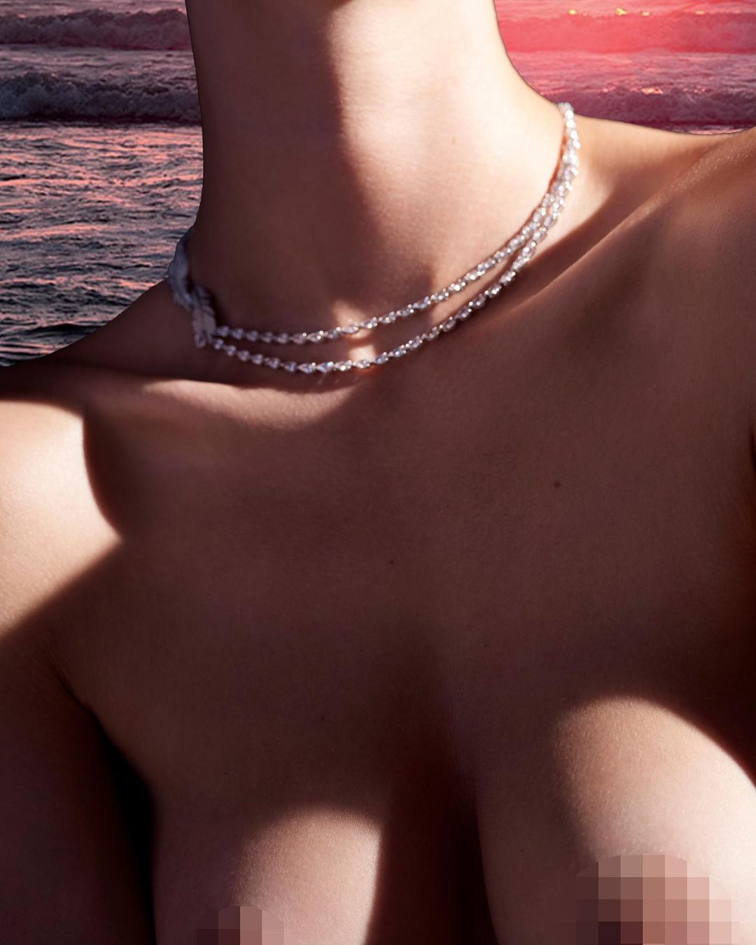 Woman with jewelry on a beach at sunset. Necklace, shoulders, and pink-tinged ocean waves visible in a close-up shot.