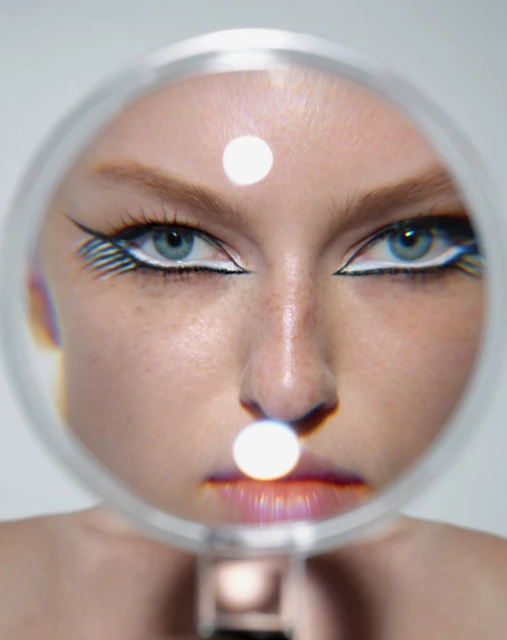 A close-up of a woman's face with bold makeup, viewed through a magnifying glass, accentuating her features in a surreal, beauty shot.