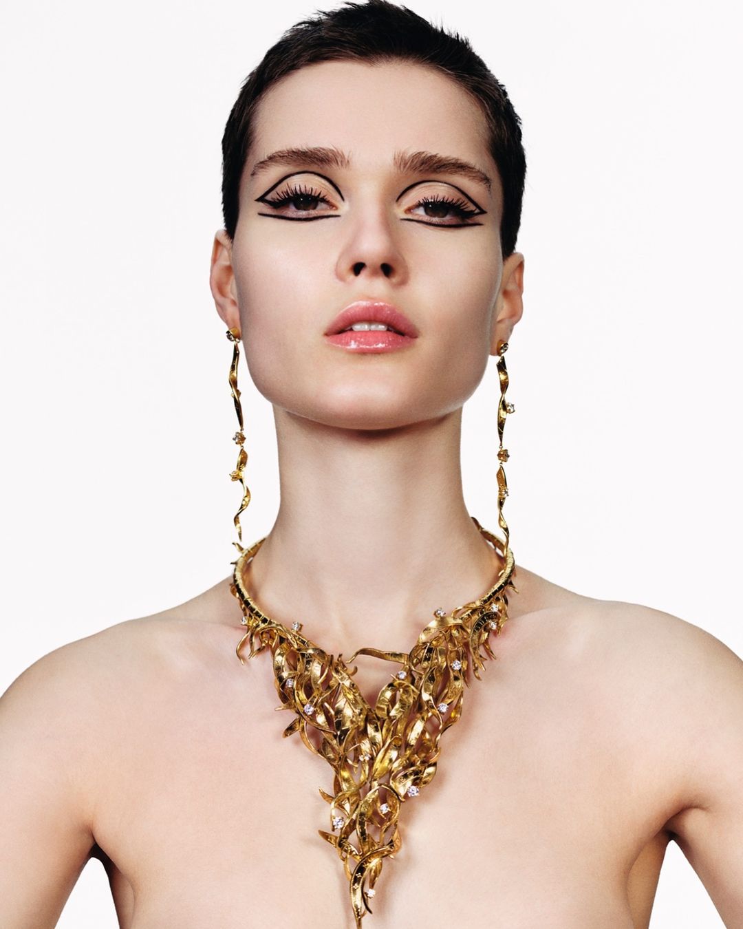 Close-up portrait of a model with short dark hair wearing a large gold statement necklace and graphic black eyeliner against a plain white background.
