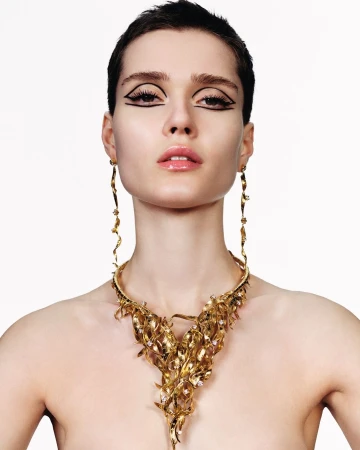 Close-up portrait of a model with short dark hair wearing a large gold statement necklace and graphic black eyeliner against a plain white background.