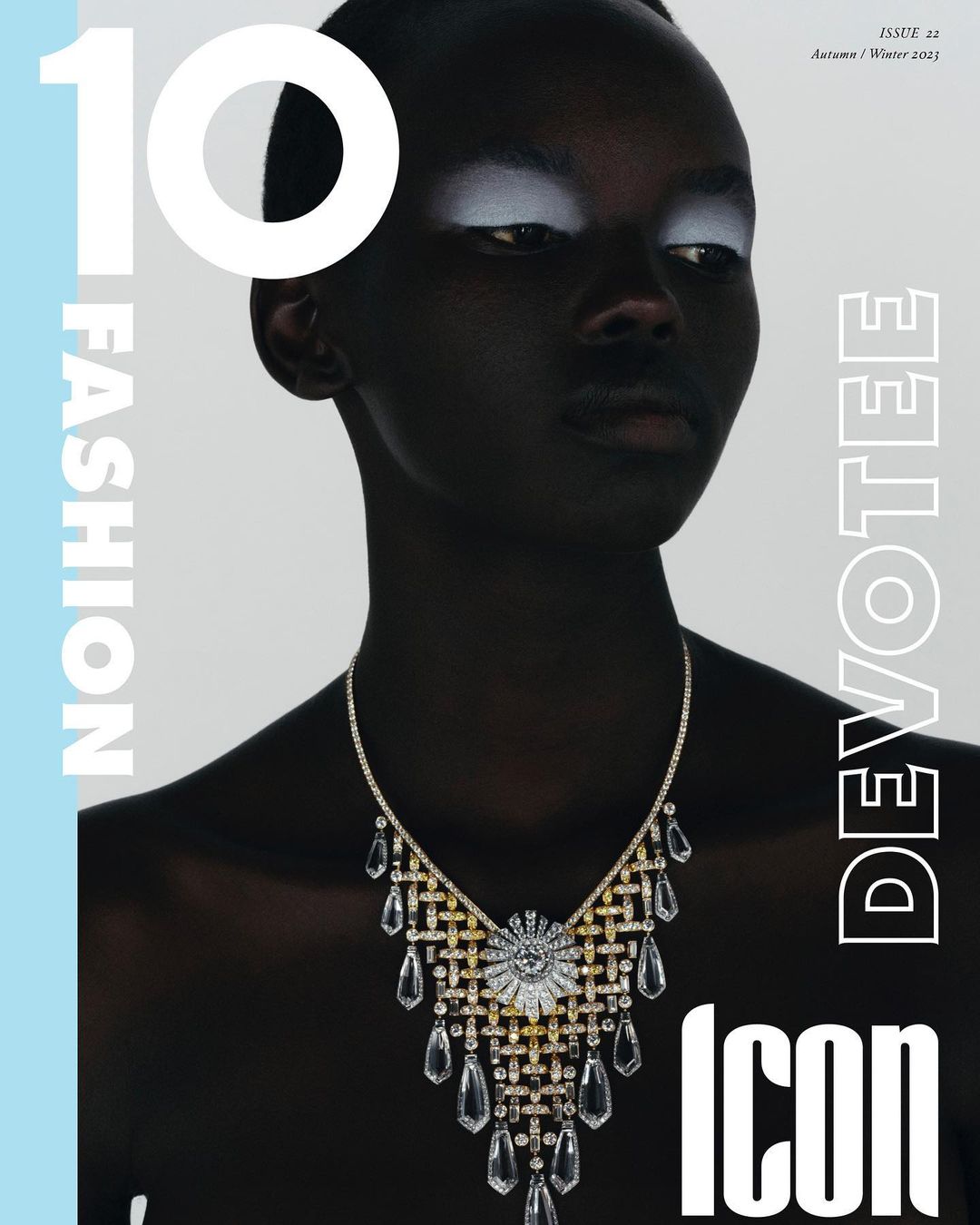 Fashion magazine cover featuring a dark-skinned adult female with white eyeshadow and an elaborate crystal necklace. The background is light blue.