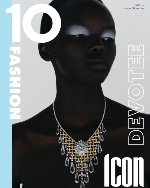 Fashion magazine cover featuring a dark-skinned adult female with white eyeshadow and an elaborate crystal necklace. The background is light blue.