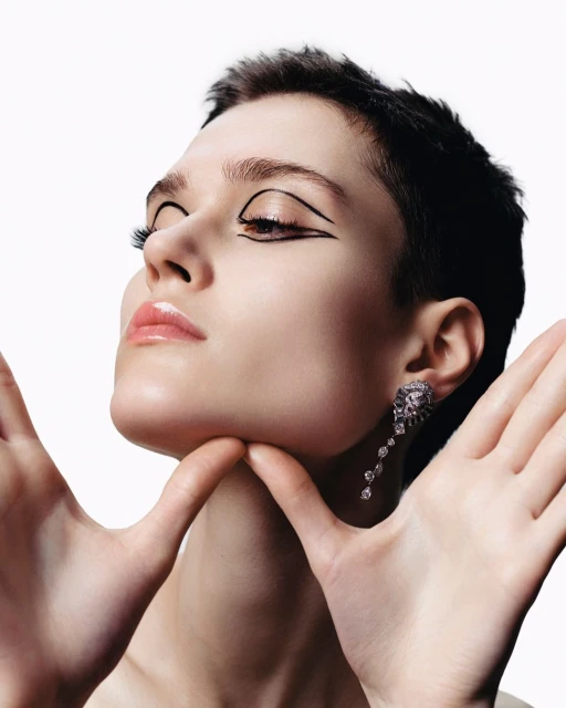 Close-up of a young adult woman with graphic eyeliner and dangling earrings, posing with hands framing her face against a white backdrop.