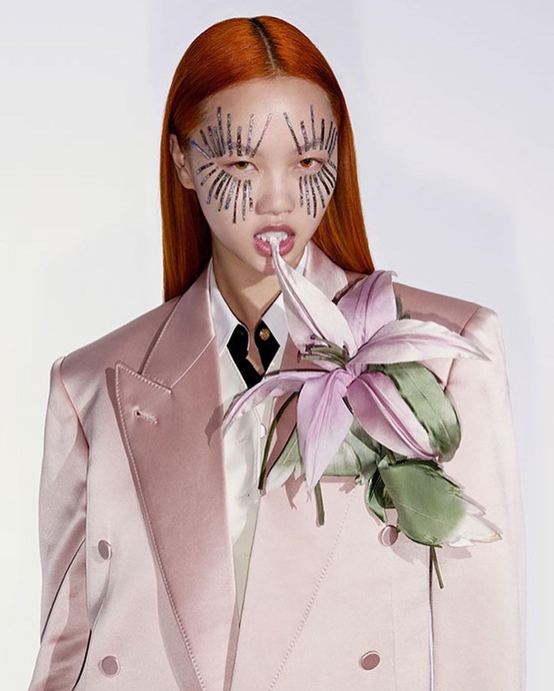 A model with orange hair and face embellishments poses in a pink blazer with a flower on a white background in a studio shot.