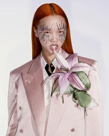 A model with orange hair and face embellishments poses in a pink blazer with a flower on a white background in a studio shot.