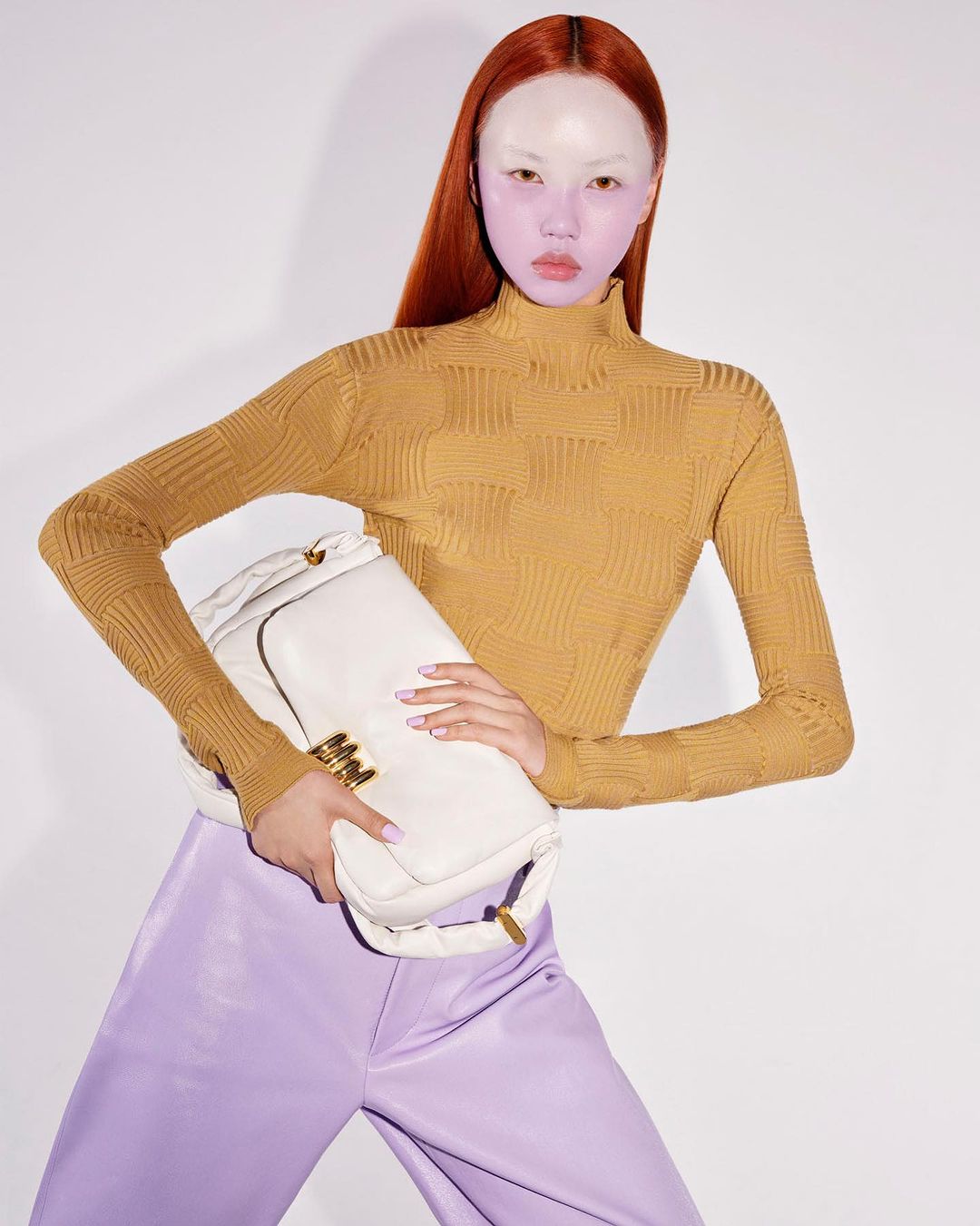 Woman poses with a large white purse in a studio setting, wearing a gold turtleneck and lavender satin pants, against a white backdrop.