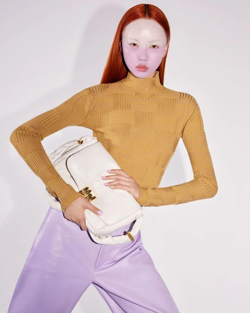 Woman poses with a large white purse in a studio setting, wearing a gold turtleneck and lavender satin pants, against a white backdrop.