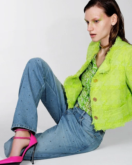 A fashion shot of a model sitting indoors wearing a green jacket, matching green top, blue jeans, and pink high heels against a white background.