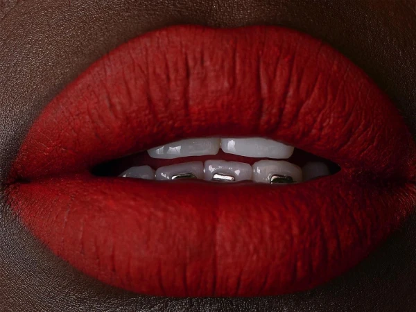Close-up macro shot of dark-skinned lips wearing bold matte red lipstick with white teeth and metallic braces visible