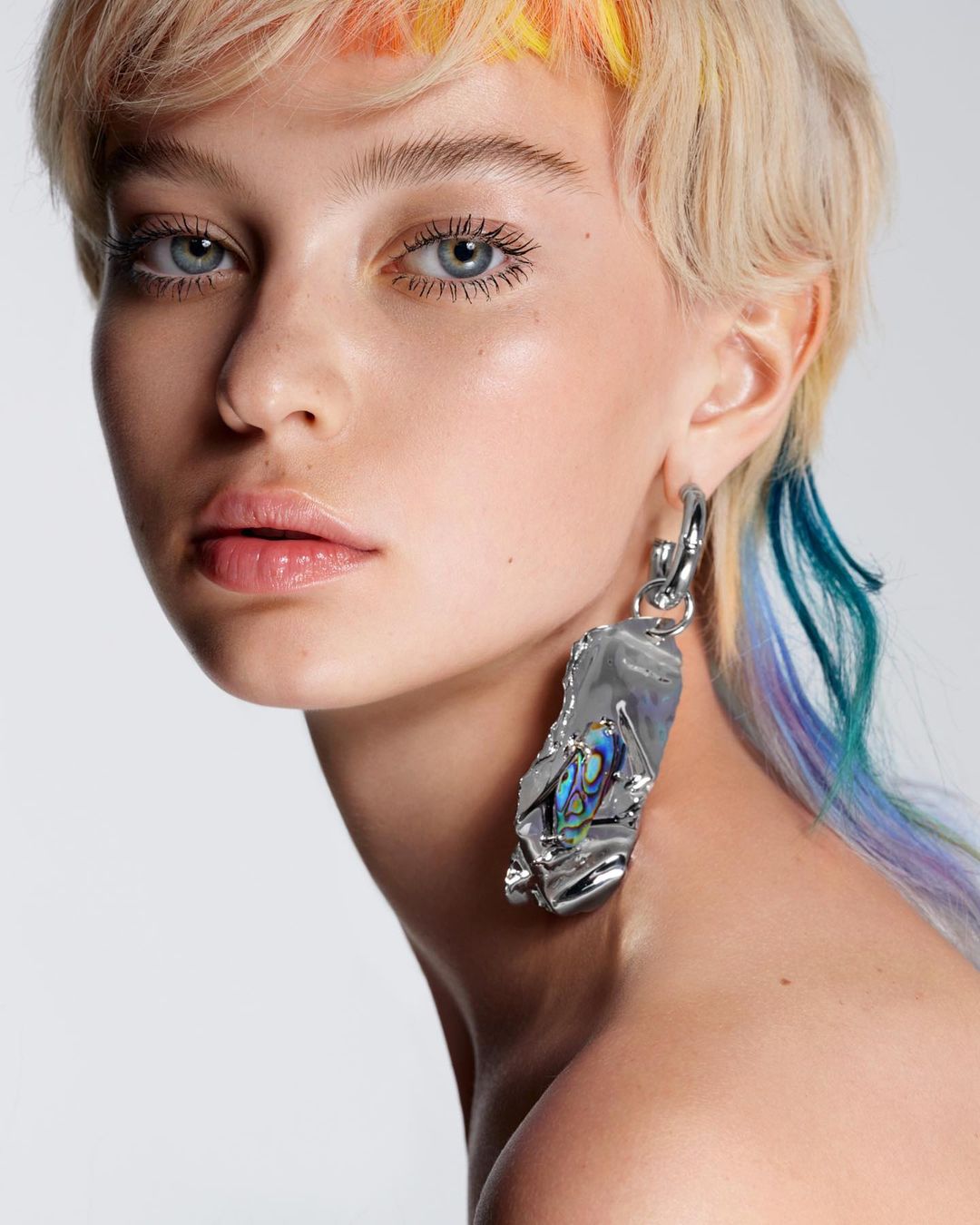 Close-up portrait of a young fair-skinned woman with dyed blonde hair wearing a large abstract silver earring against a light gray backdrop.